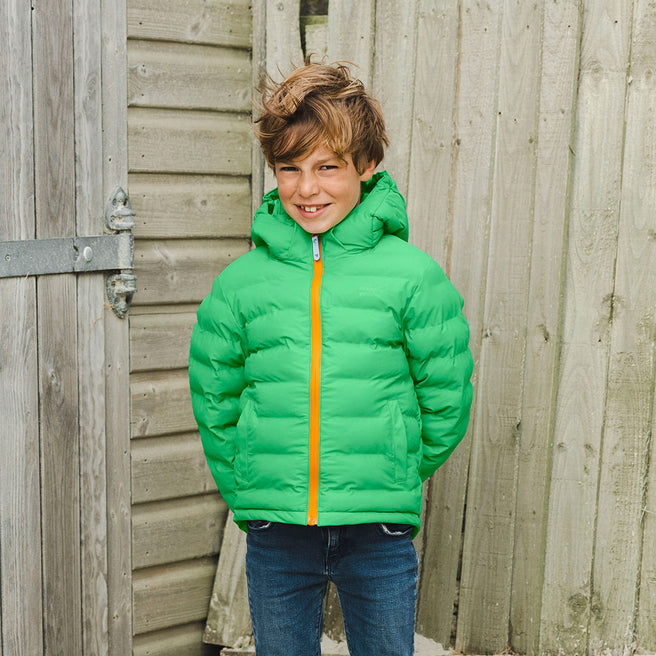 A boy in a Waterproof Puffer Jacket Green smiles, standing in front of a wooden door and fence.